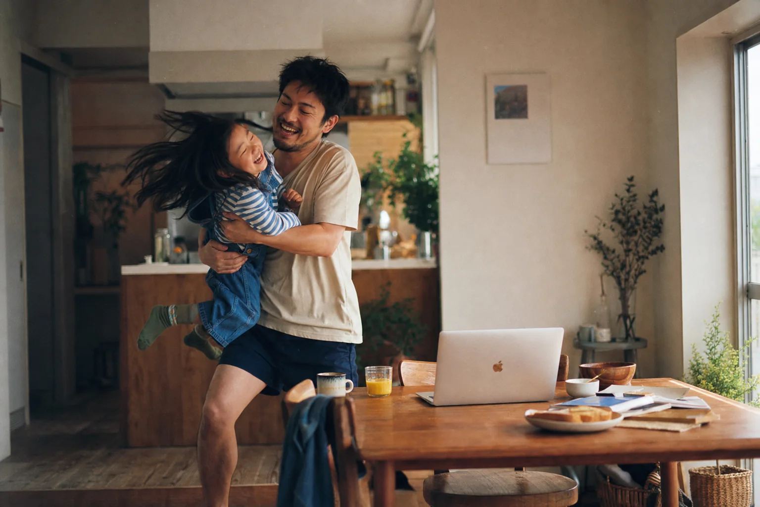 A father laughing as he lifts his daughter in a bright kitchen beside an open MacBook